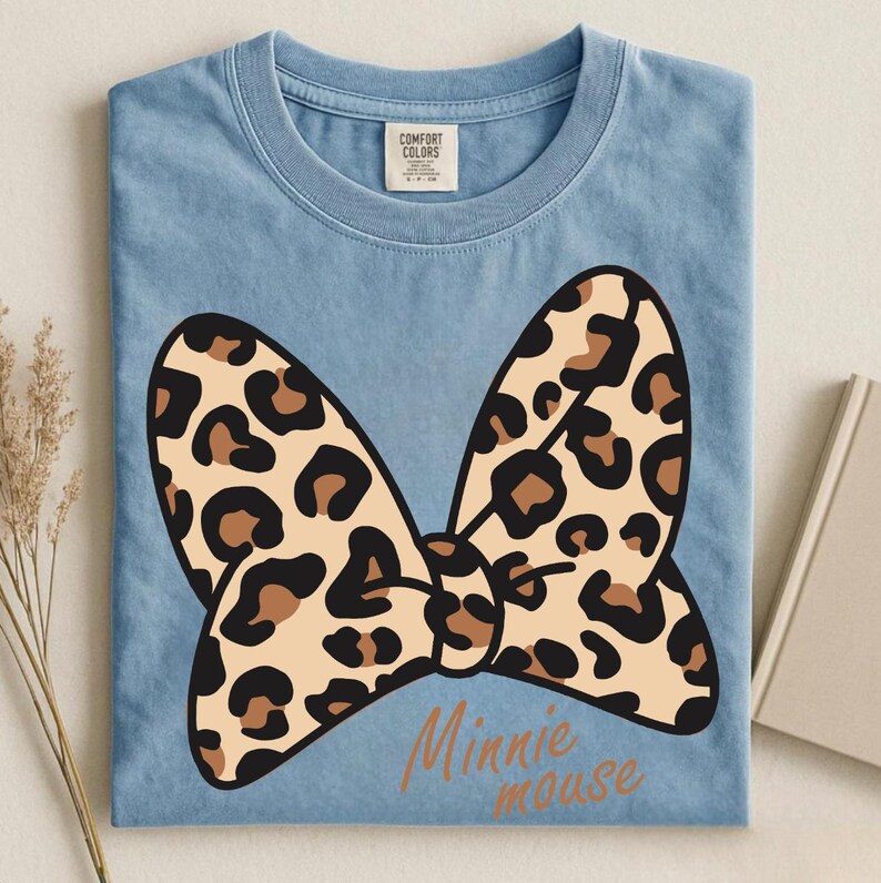 May include: Light blue t-shirt featuring a large bow with a leopard print design. The bow is tan with brown spots. The words "Minnie mouse" are printed in a cursive font below the bow. The shirt is folded.