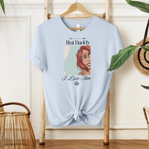 May include: Light blue t-shirt with a graphic featuring a cartoon illustration of a character with red hair. The text on the shirt reads "But Daddy I Love Him" with the year "1989" above the text. A seashell graphic is below the text.