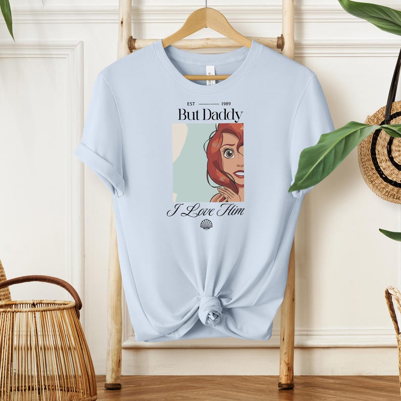 May include: Light blue t-shirt with a graphic featuring a cartoon illustration of a character with red hair. The text on the shirt reads "But Daddy I Love Him" with the year "1989" above the text. A seashell graphic is below the text.