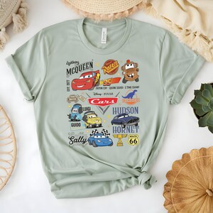 May include: A light green t-shirt featuring a colorful graphic of characters from the movie "Cars." The design includes Lightning McQueen, Mater, and other characters, along with the text "Cars" and "Route 66."