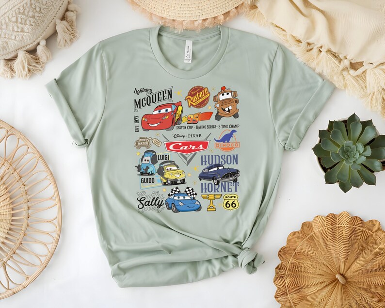 May include: A light green t-shirt featuring a colorful graphic of characters from the movie "Cars." The design includes Lightning McQueen, Mater, and other characters, along with the text "Cars" and "Route 66."