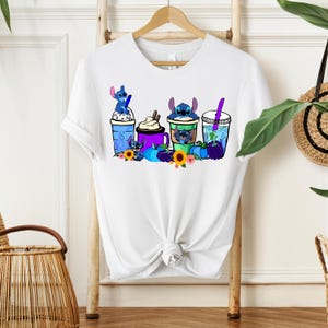 Puede incluir: Camiseta blanca con un gr&aacute;fico colorido de personajes de Stitch con bebidas, calabazas y flores. El dise&ntilde;o incluye elementos azules, morados y amarillos. La camiseta est&aacute; colgada en un perchero de madera.