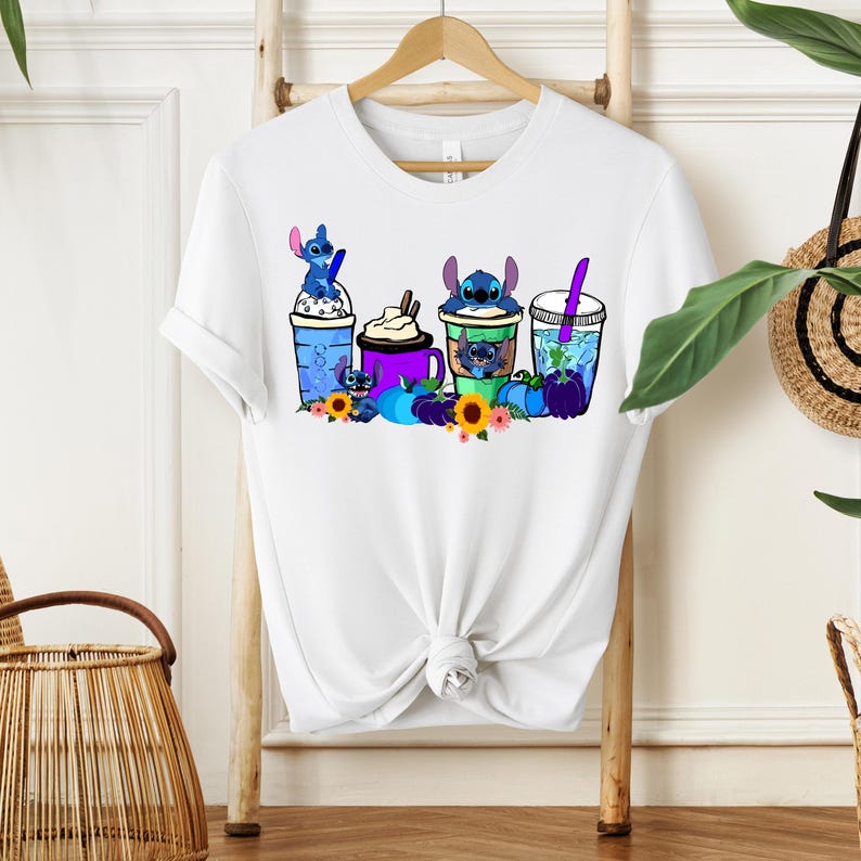 Puede incluir: Camiseta blanca con un gr&aacute;fico colorido de personajes de Stitch con bebidas, calabazas y flores. El dise&ntilde;o incluye elementos azules, morados y amarillos. La camiseta est&aacute; colgada en un perchero de madera.