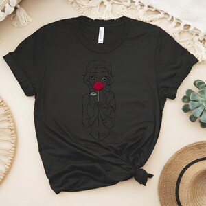 May include: Black t-shirt featuring a line drawing of a person holding a red rose. The design is a simple outline with the rose in full color. The shirt is made of a soft material and has short sleeves.