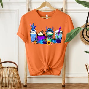 Puede incluir: Camiseta naranja con un dise&ntilde;o de personajes de Stitch en varias bebidas. El dise&ntilde;o incluye bebidas azules, moradas y verdes con personajes de Stitch, girasoles y calabazas. La camiseta est&aacute; colgada en una percha de madera.