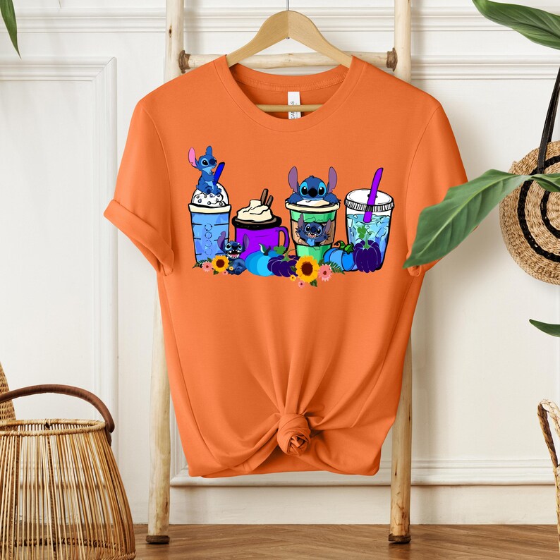 Puede incluir: Camiseta naranja con un dise&ntilde;o de personajes de Stitch en varias bebidas. El dise&ntilde;o incluye bebidas azules, moradas y verdes con personajes de Stitch, girasoles y calabazas. La camiseta est&aacute; colgada en una percha de madera.