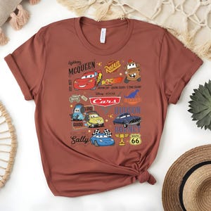 May include: A rust-colored t-shirt featuring a graphic design of characters from the animated movie "Cars." The design includes Lightning McQueen, Mater, and other characters, along with the word "Cars" and other text.