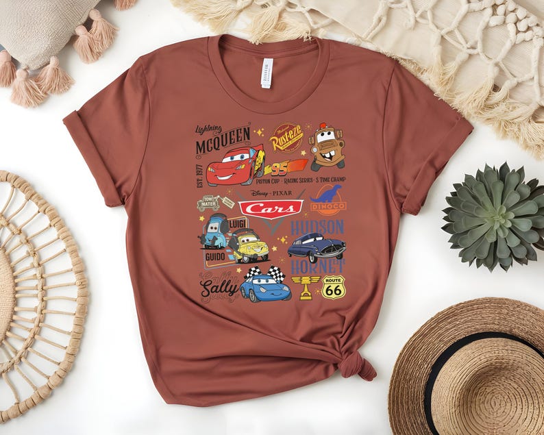 May include: A rust-colored t-shirt featuring a graphic design of characters from the animated movie "Cars." The design includes Lightning McQueen, Mater, and other characters, along with the word "Cars" and other text.