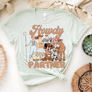 May include: A light green t-shirt featuring a cartoon graphic with the text "Howdy Partner". The graphic includes characters from a popular animated movie, including a cowboy, a cowgirl, and a space ranger.
