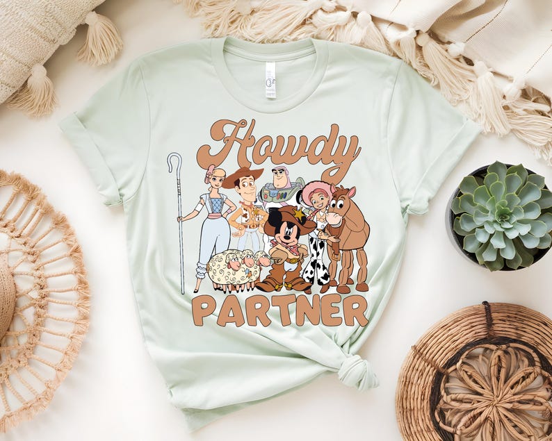 May include: A light green t-shirt featuring a cartoon graphic with the text "Howdy Partner". The graphic includes characters from a popular animated movie, including a cowboy, a cowgirl, and a space ranger.
