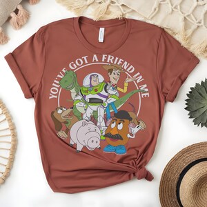 May include: A terracotta-coloured t-shirt with a graphic of characters from the animated film Toy Story. The design includes Woody, Buzz Lightyear, Rex, Slinky Dog, Hamm, and Mr. Potato Head, with the text "You've got a friend in me" in a circular design.