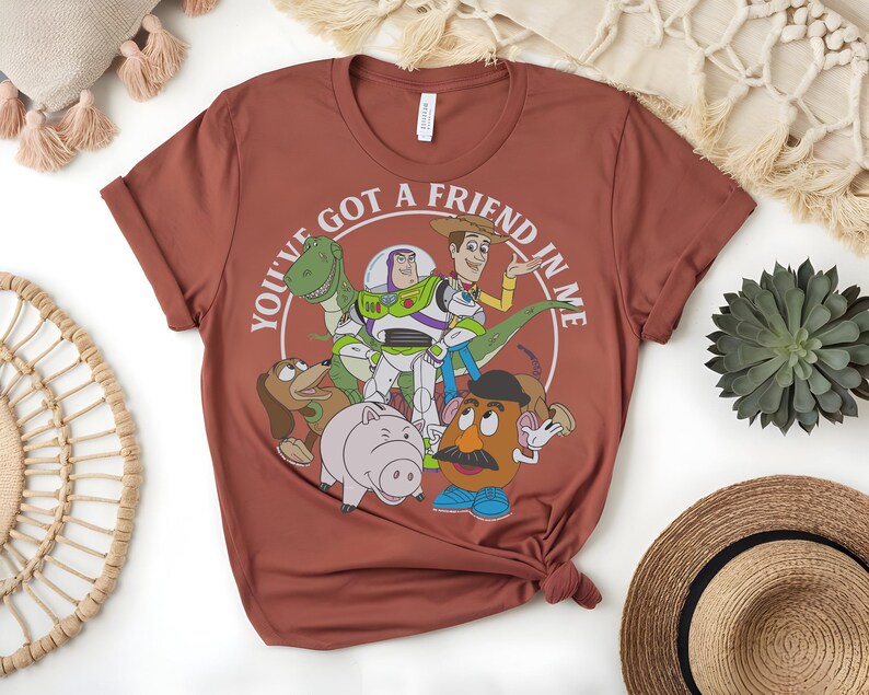 May include: A terracotta-coloured t-shirt with a graphic of characters from the animated film Toy Story. The design includes Woody, Buzz Lightyear, Rex, Slinky Dog, Hamm, and Mr. Potato Head, with the text "You've got a friend in me" in a circular design.