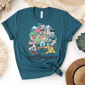 Puede incluir: Camiseta de color turquesa con un gr&aacute;fico colorido de personajes de Disney, incluyendo Mickey Mouse y Donald Duck, frente a un castillo. El texto "Walt Disney World" est&aacute; impreso debajo.