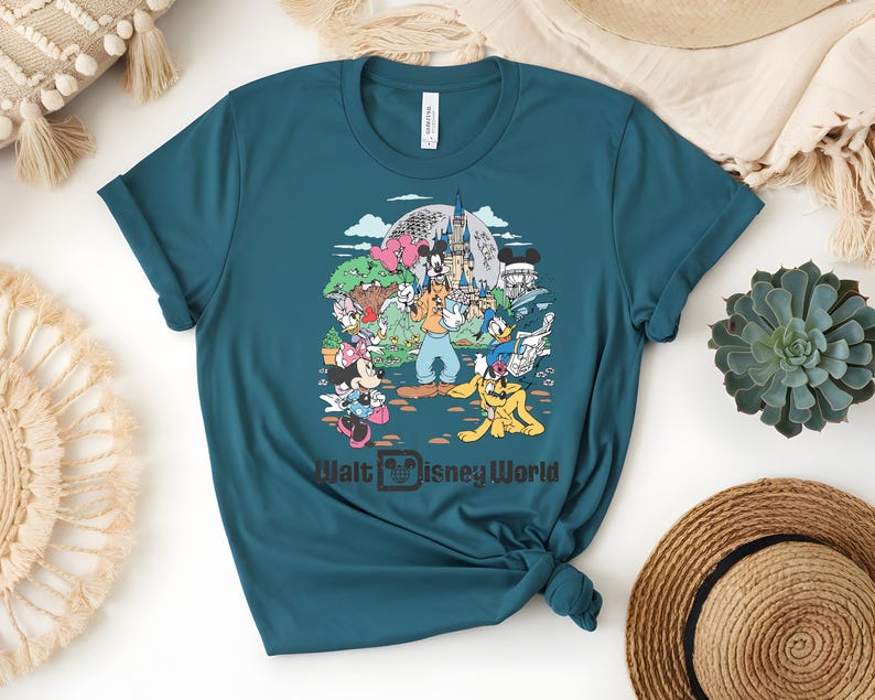 Puede incluir: Camiseta de color turquesa con un gr&aacute;fico colorido de personajes de Disney, incluyendo Mickey Mouse y Donald Duck, frente a un castillo. El texto "Walt Disney World" est&aacute; impreso debajo.
