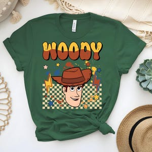 Puede incluir: Camiseta verde oscuro con la palabra "WOODY" en letras retro naranjas y marrones. La camiseta presenta una imagen de dibujos animados de un vaquero con un sombrero marr&oacute;n, sobre un fondo a cuadros con estrellas y rayos.