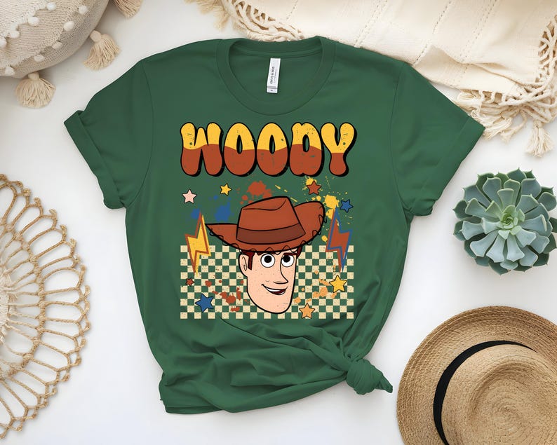 Puede incluir: Camiseta verde oscuro con la palabra "WOODY" en letras retro naranjas y marrones. La camiseta presenta una imagen de dibujos animados de un vaquero con un sombrero marr&oacute;n, sobre un fondo a cuadros con estrellas y rayos.