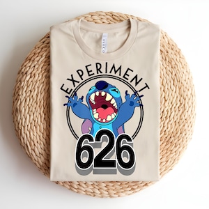 Pu&ograve; includere: Maglietta beige con un personaggio dei cartoni animati blu con la bocca aperta e il numero "626". Il personaggio &egrave; all'interno di un cerchio nero con la scritta "EXPERIMENT" sopra. La maglietta &egrave; piegata su un tappetino intrecciato.