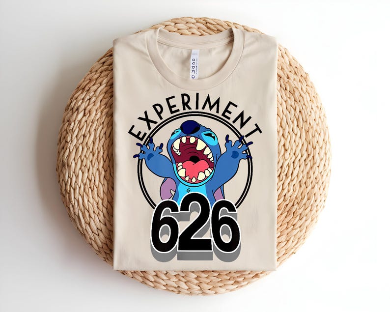 Pu&ograve; includere: Maglietta beige con un personaggio dei cartoni animati blu con la bocca aperta e il numero "626". Il personaggio &egrave; all'interno di un cerchio nero con la scritta "EXPERIMENT" sopra. La maglietta &egrave; piegata su un tappetino intrecciato.