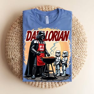 May include: A light blue t-shirt with a graphic of Darth Vader grilling with two Stormtroopers. The text "Darthlorian" is above the image, and Darth Vader wears a red apron that says "#1 Dad". The shirt is folded on a woven surface.