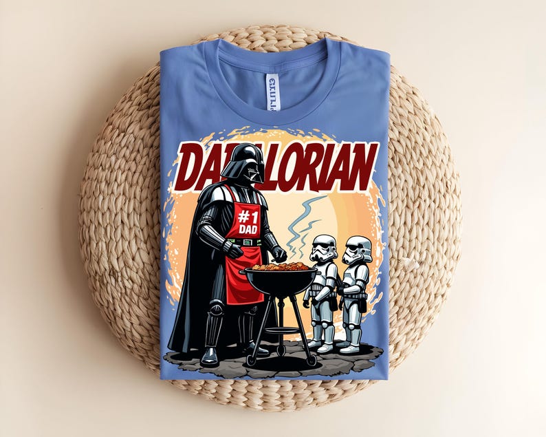 May include: A light blue t-shirt with a graphic of Darth Vader grilling with two Stormtroopers. The text "Darthlorian" is above the image, and Darth Vader wears a red apron that says "#1 Dad". The shirt is folded on a woven surface.