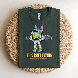 May include: Dark green t-shirt featuring a cartoon character in a Buzz Lightyear costume with the text "THIS ISN'T FLYING" and "This is falling with style." The character is blue with large eyes and a pink ear. The shirt is on a woven surface.