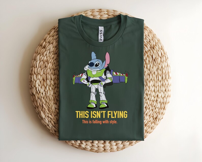 May include: Dark green t-shirt featuring a cartoon character in a Buzz Lightyear costume with the text "THIS ISN'T FLYING" and "This is falling with style." The character is blue with large eyes and a pink ear. The shirt is on a woven surface.