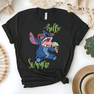 K&ouml;nnte beinhalten: Schwarzes T-Shirt mit einer Comicfigur, die eine Sonnenbrille tr&auml;gt und eine Eist&uuml;te h&auml;lt. Die Worte "hello summer" sind in gr&uuml;ner Schrift &uuml;ber und unter der Figur geschrieben.