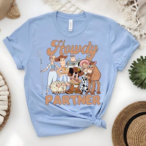 May include: Light blue t-shirt featuring a cartoon graphic of characters with the text "Howdy Partner" in a retro font. The graphic includes a sheep, a cowboy, a horse, and other characters. The shirt is made of a soft material.