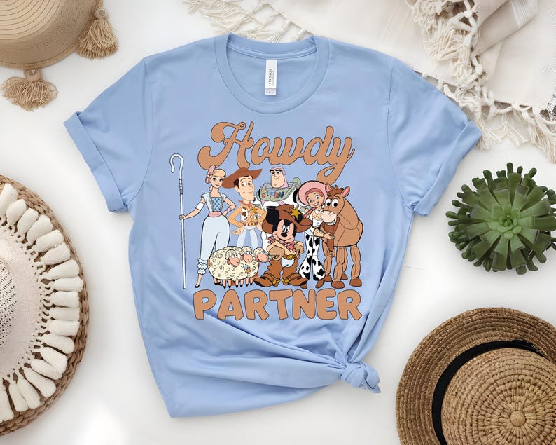 May include: Light blue t-shirt featuring a cartoon graphic of characters with the text "Howdy Partner" in a retro font. The graphic includes a sheep, a cowboy, a horse, and other characters. The shirt is made of a soft material.