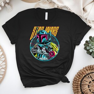 May include: Black t-shirt with a vibrant graphic of a Star Wars character in a circular frame. The character, wearing a helmet and armour, is holding a weapon. The words "STAR WARS" are above the character in yellow and orange.