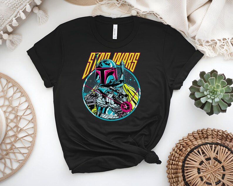 May include: Black t-shirt with a vibrant graphic of a Star Wars character in a circular frame. The character, wearing a helmet and armour, is holding a weapon. The words "STAR WARS" are above the character in yellow and orange.