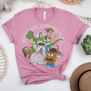 May include: A pink t-shirt with a graphic of characters from the animated film "Toy Story." The design includes Woody, Buzz Lightyear, Rex, Slinky Dog, Hamm, and Mr. Potato Head, with the text "YOU'VE GOT A FRIEND IN ME" encircling the characters.