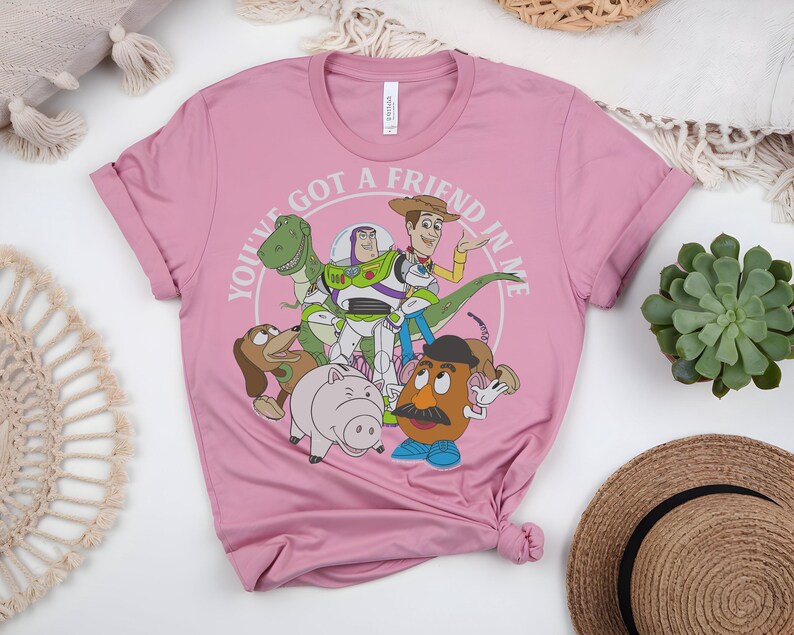 May include: A pink t-shirt with a graphic of characters from the animated film "Toy Story." The design includes Woody, Buzz Lightyear, Rex, Slinky Dog, Hamm, and Mr. Potato Head, with the text "YOU'VE GOT A FRIEND IN ME" encircling the characters.