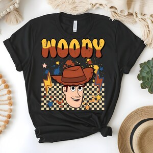 Puede incluir: Camiseta negra con un dise&ntilde;o retro que presenta el nombre "WOODY" en letras marrones y amarillas. El gr&aacute;fico incluye un personaje de dibujos animados de vaquero con un sombrero marr&oacute;n, sobre un fondo a cuadros con estrellas y rayos.