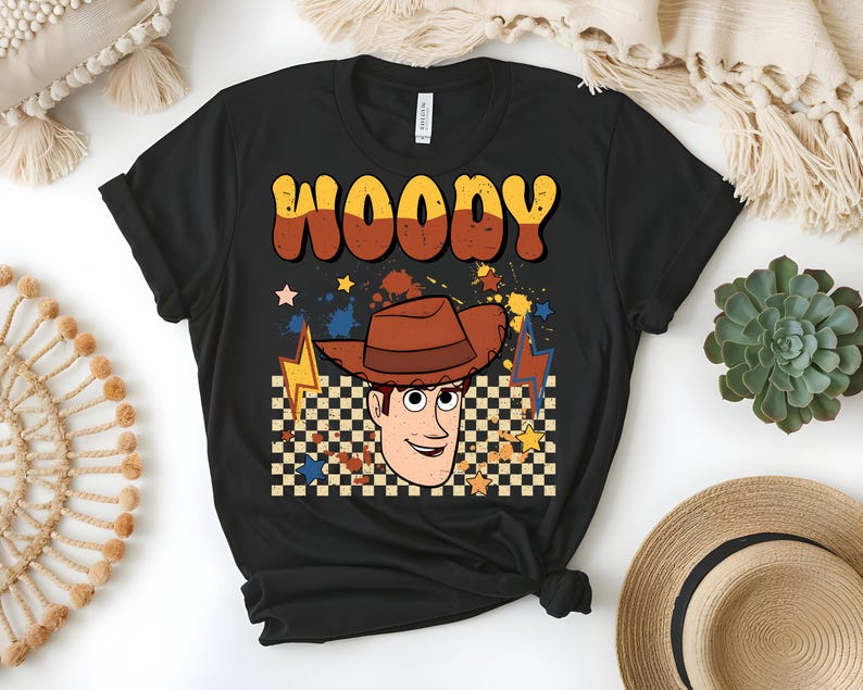 Puede incluir: Camiseta negra con un dise&ntilde;o retro que presenta el nombre "WOODY" en letras marrones y amarillas. El gr&aacute;fico incluye un personaje de dibujos animados de vaquero con un sombrero marr&oacute;n, sobre un fondo a cuadros con estrellas y rayos.