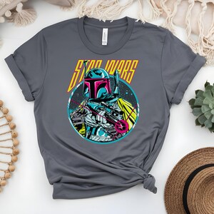 May include: A grey t-shirt with a graphic of a Star Wars character. The design features a teal, pink, and yellow helmeted figure against a starry backdrop, with the words "STAR WARS" in yellow.