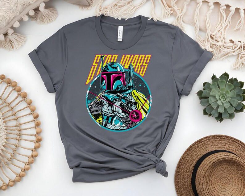 May include: A grey t-shirt with a graphic of a Star Wars character. The design features a teal, pink, and yellow helmeted figure against a starry backdrop, with the words "STAR WARS" in yellow.