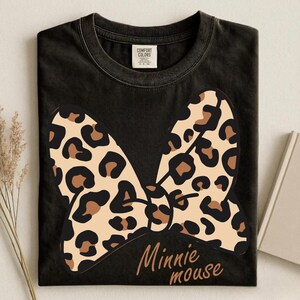 May include: Black t-shirt featuring a large bow with a leopard print design in tan, brown, and black. The words "Minnie mouse" are written in a brown script font below the bow. The shirt is from the Comfort Colors brand.