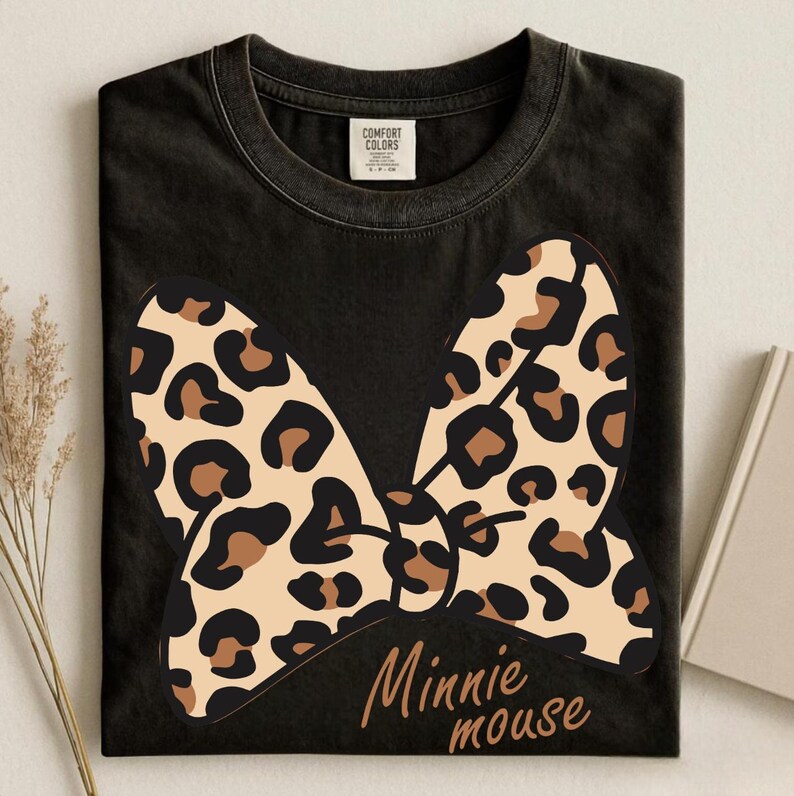 May include: Black t-shirt featuring a large bow with a leopard print design in tan, brown, and black. The words "Minnie mouse" are written in a brown script font below the bow. The shirt is from the Comfort Colors brand.