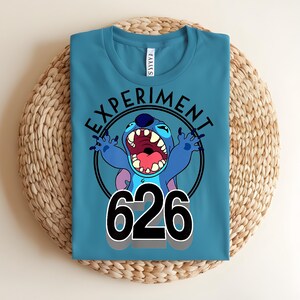 Pu&ograve; includere: T-shirt color teal con un'immagine a cartoni animati di Stitch, le parole "EXPERIMENT" e il numero "626". La grafica &egrave; in un cerchio nero. La maglietta &egrave; piegata su una superficie circolare intrecciata.