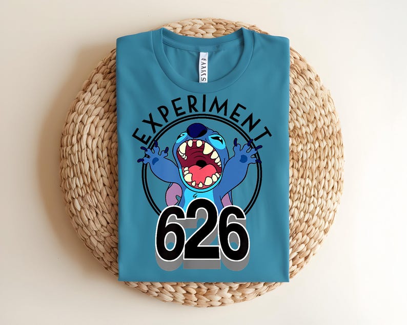 Pu&ograve; includere: T-shirt color teal con un'immagine a cartoni animati di Stitch, le parole "EXPERIMENT" e il numero "626". La grafica &egrave; in un cerchio nero. La maglietta &egrave; piegata su una superficie circolare intrecciata.