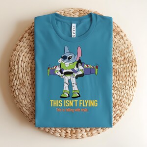 May include: Teal t-shirt featuring a cartoon character, Stitch, dressed as Buzz Lightyear with the text "THIS ISN'T FLYING" and "This is falling with style." The shirt is folded on a woven, circular surface.