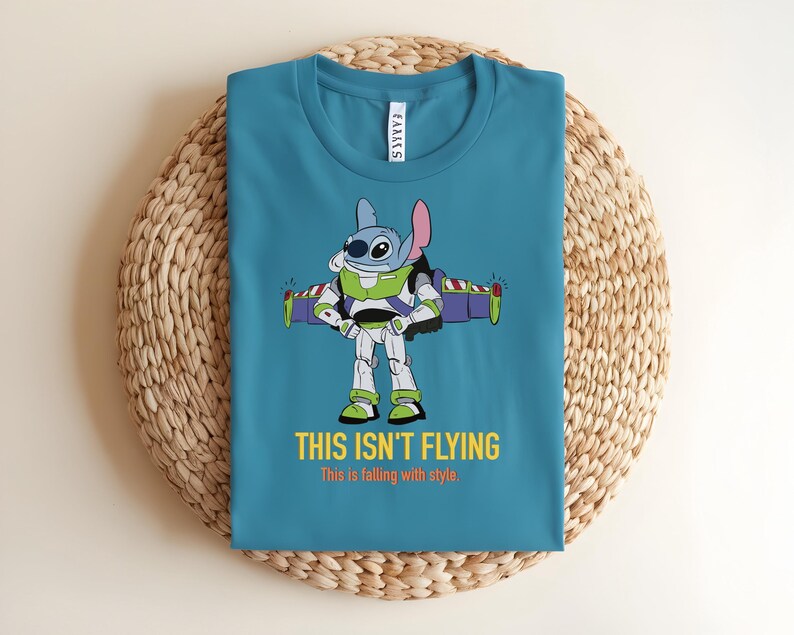 May include: Teal t-shirt featuring a cartoon character, Stitch, dressed as Buzz Lightyear with the text "THIS ISN'T FLYING" and "This is falling with style." The shirt is folded on a woven, circular surface.
