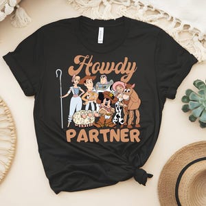 May include: Black t-shirt featuring a cartoon graphic of characters from a popular animated movie. The text "Howdy Partner" is printed in a brown, western-style font. The design includes a sheep, a horse, and other characters.