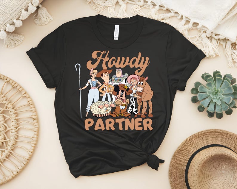 May include: Black t-shirt featuring a cartoon graphic of characters from a popular animated movie. The text "Howdy Partner" is printed in a brown, western-style font. The design includes a sheep, a horse, and other characters.