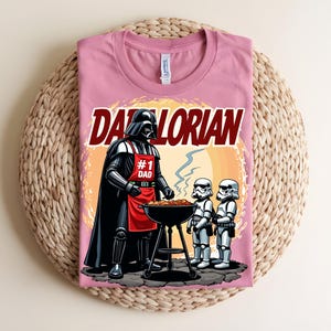 May include: A pink t-shirt with a graphic of Darth Vader grilling with Stormtroopers. The text "Darthlorian" is above the image. Darth Vader wears a red apron that says "#1 Dad". The shirt is on a woven surface.