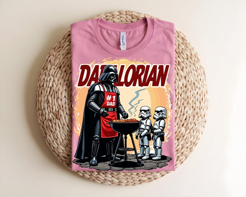May include: A pink t-shirt with a graphic of Darth Vader grilling with Stormtroopers. The text "Darthlorian" is above the image. Darth Vader wears a red apron that says "#1 Dad". The shirt is on a woven surface.
