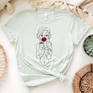 May include: Light green t-shirt featuring a black and white line drawing of a woman holding a red rose to her face. The woman has long wavy hair and is wearing a collared shirt. The t-shirt is knotted at the bottom.