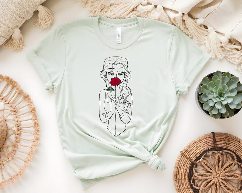 May include: Light green t-shirt featuring a black and white line drawing of a woman holding a red rose to her face. The woman has long wavy hair and is wearing a collared shirt. The t-shirt is knotted at the bottom.
