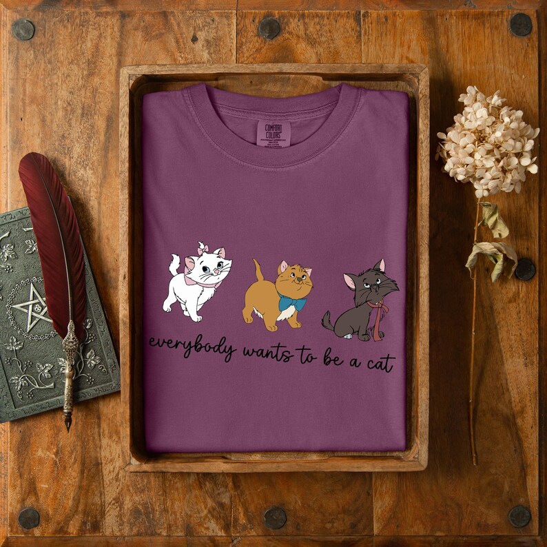 May include: A plum-coloured t-shirt with three cartoon cats, each with a bow or scarf. The phrase "everybody wants to be a cat" is printed below. The shirt is presented in a wooden tray.